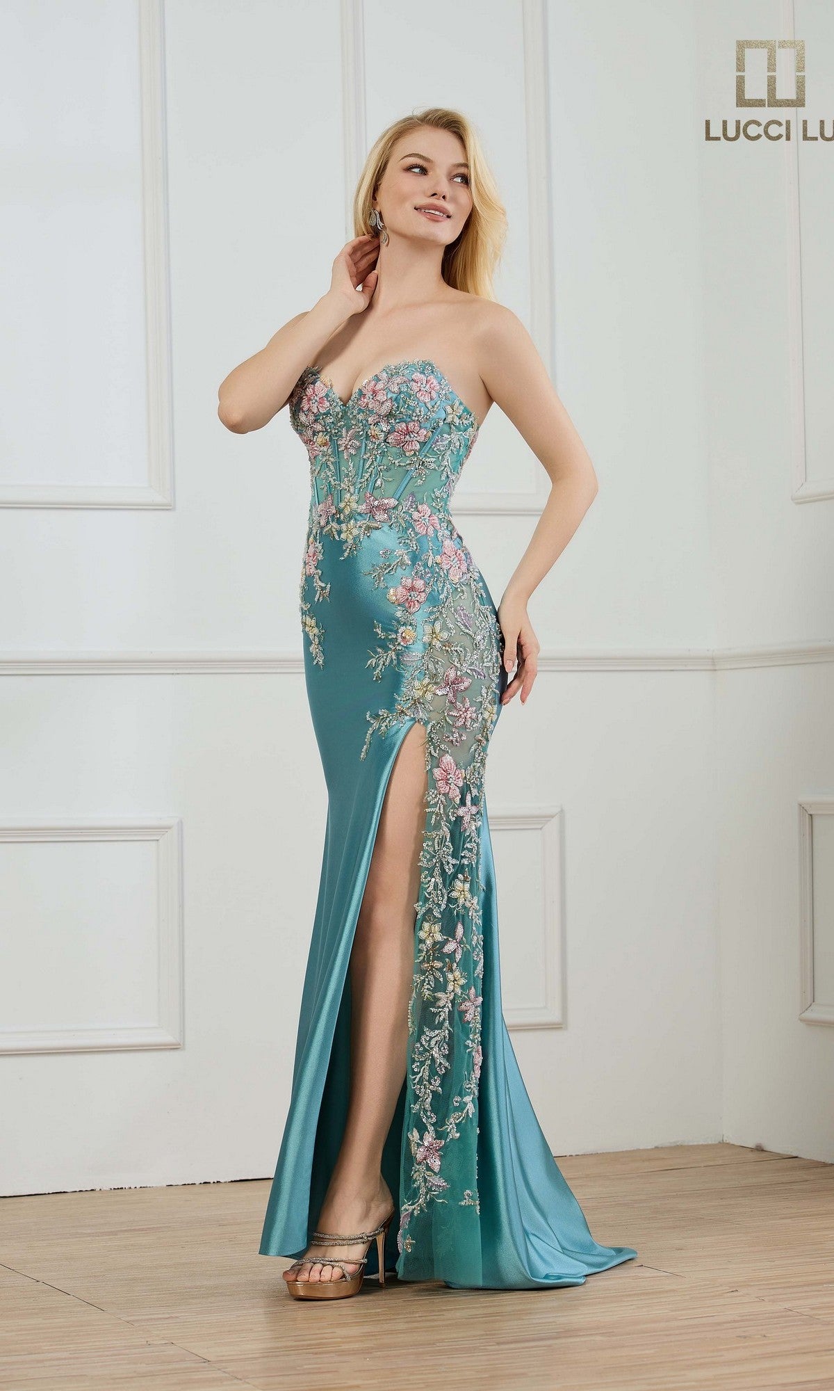 A woman stands in a white room with wood flooring, wearing the Lucci Lu 1435 long teal prom dress in stretch satin with floral embroidery, a strapless neckline, high slit, and high-heeled sandals.